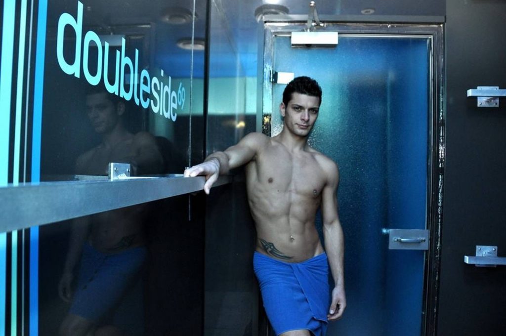 Un homosexuel en maillot de bain bleu devant une porte à Lyon.